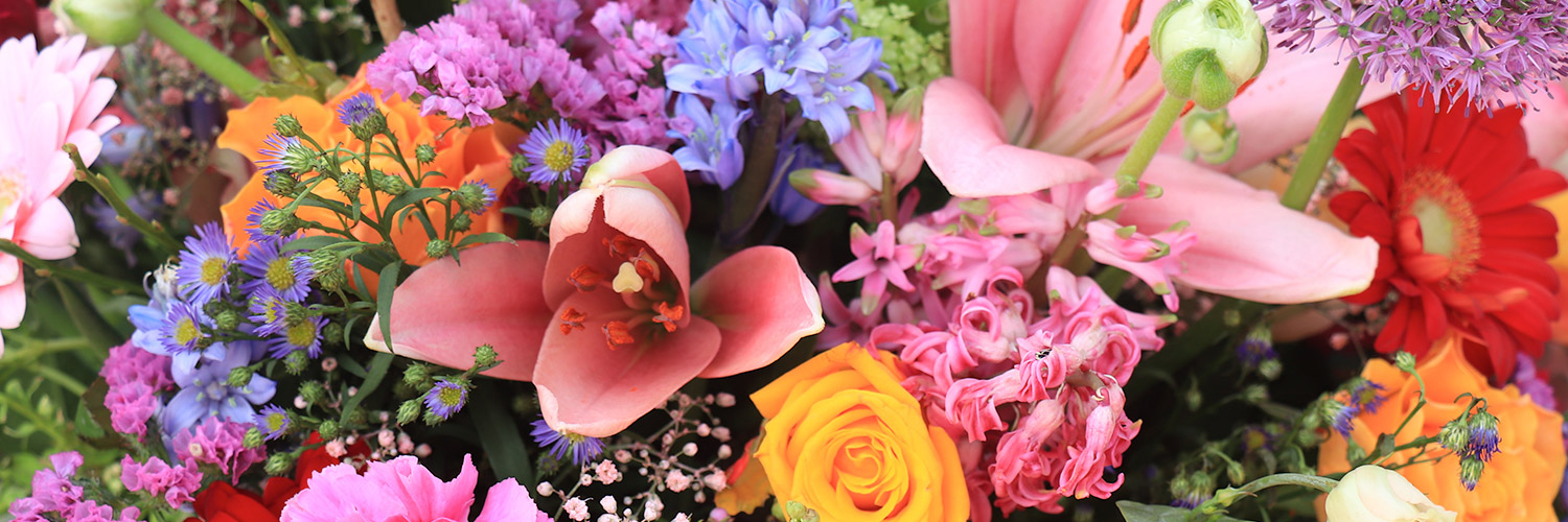 A vibrant bouquet displays various colorful flowers including lilies, roses, and daisies, surrounded by green foliage and small blue, purple, and pink blossoms, creating a lush, visually striking arrangement.