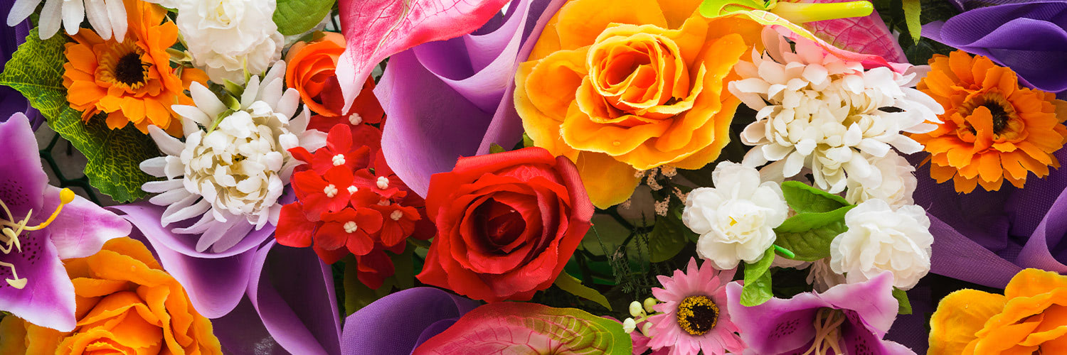 Vibrant flowers in assorted colors including red, orange, white, and purple are arranged together, surrounded by green leaves and purple ribbon, creating a visually stunning bouquet.